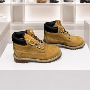 May include: A pair of tan nubuck leather boots featuring a black padded collar, gold-tone metal eyelets, and yellow and brown woven laces. The boots have a thick, dark brown rubber lug sole and a small embossed tree logo on the side heel area.