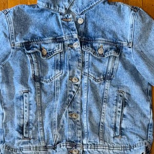 May include: Pale blue denim jacket with a classic design. Features a collared neckline, button-down front, two chest pockets, and two side pockets. The jacket has a slightly faded wash and silver-tone buttons. A casual outerwear piece.