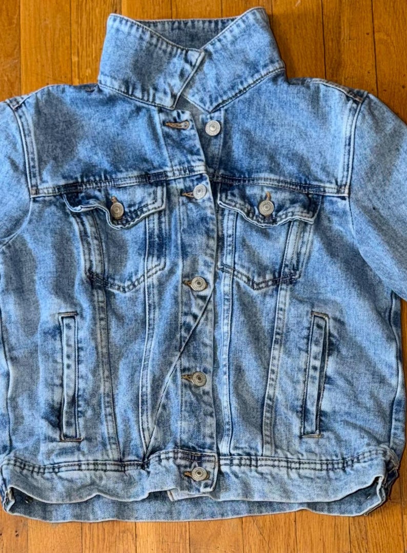 May include: Pale blue denim jacket with a classic design. Features a collared neckline, button-down front, two chest pockets, and two side pockets. The jacket has a slightly faded wash and silver-tone buttons. A casual outerwear piece.