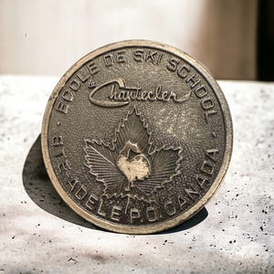 May include: A round, bronze-colored metal medallion with the text "ECOLE DE SKI SCHOOL" and "STE ADELE P.O. CANADA" around the edge. The center features a maple leaf with a stylized bird. The word "Chantecler" is in cursive.