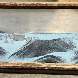 May include: A rectangular sand art piece with a wooden frame. The artwork features a landscape with black and gray mountains, a light blue sky, and a light blue foreground. The frame is made of light brown wood.