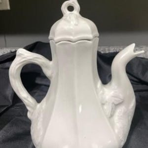 以下が含まれることがあります： 装飾的な花の模様が施された白い陶器製のティーポット。ティーポットは、上にノブが付いた蓋と、側面に注ぎ口が付いています。