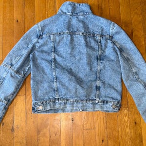 May include: A light blue denim jacket with a classic design. The jacket has a button-up front, long sleeves, and a collared neckline. The denim has a faded, acid-wash appearance. The jacket is laid flat on a wooden surface.