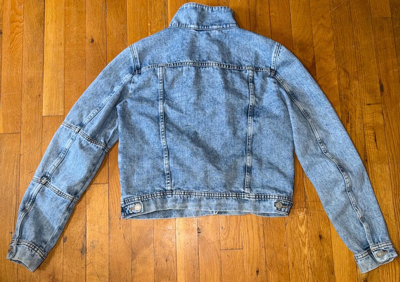 May include: A light blue denim jacket with a classic design. The jacket has a button-up front, long sleeves, and a collared neckline. The denim has a faded, acid-wash appearance. The jacket is laid flat on a wooden surface.
