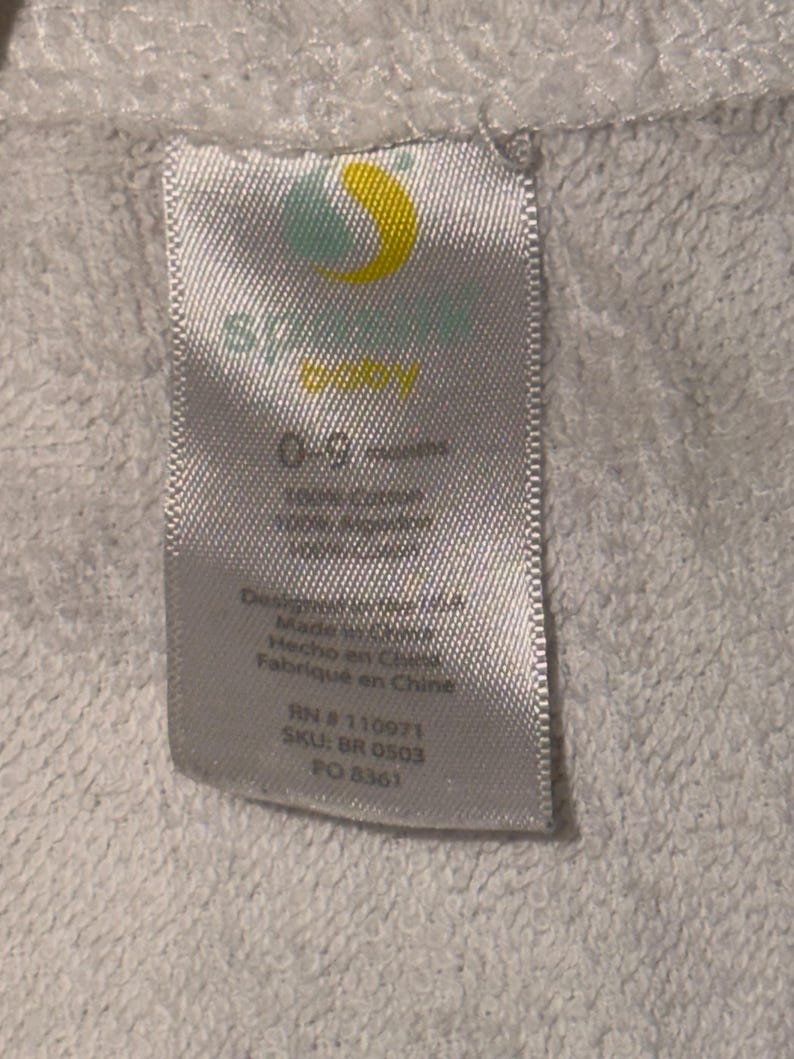 May include: A white fabric tag with the brand logo and text. The tag includes the text "0-9 months", "100% cotton", and "100% acrylic". Additional text includes "Designed in USA", "Made in China", "Hecho en China", and "Fabriqu&eacute; en Chine".