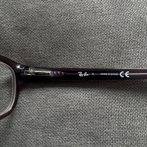 May include: Close-up of a pair of Ray-Ban eyeglasses. The frames are a dark purple color. The Ray-Ban logo and "MADE IN CHINA" text are visible on the temple arm. The glasses are resting on a gray textured surface.