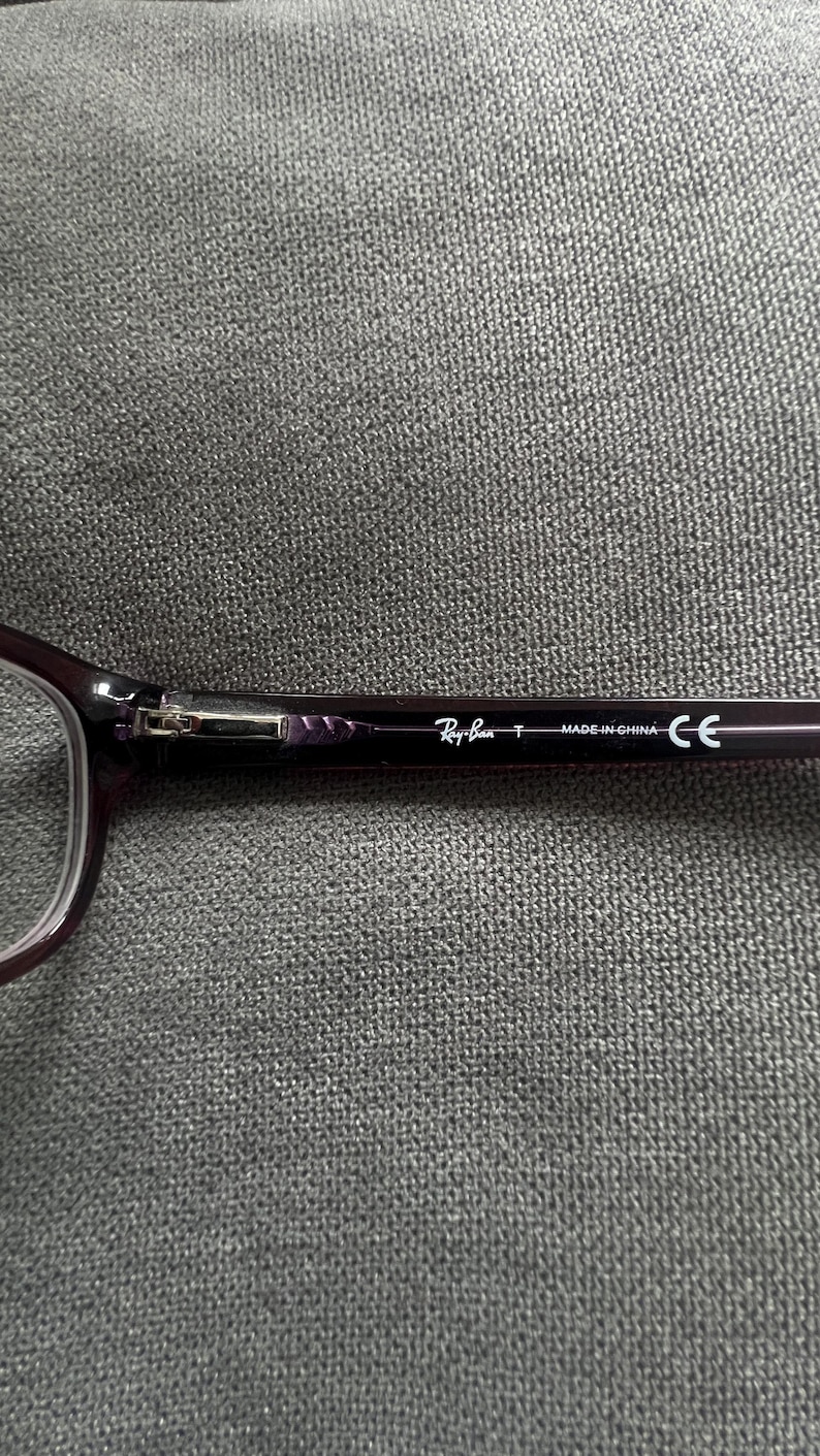 May include: Close-up of a pair of Ray-Ban eyeglasses. The frames are a dark purple color. The Ray-Ban logo and "MADE IN CHINA" text are visible on the temple arm. The glasses are resting on a gray textured surface.