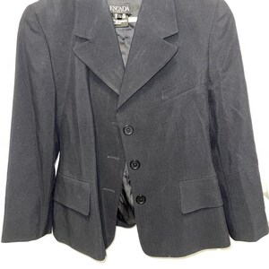 May include: A black blazer with a notched lapel, three-button closure, and two front pockets. The blazer is hanging on a wooden hanger. The label reads "ESCADA".