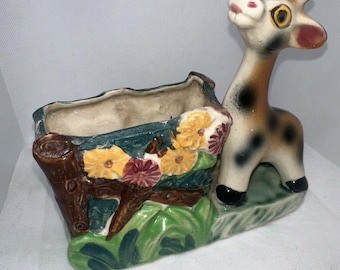 May include: A ceramic planter shaped like a giraffe with a brown, black, and white spotted pattern. The giraffe's head is turned to the side and it has a pink nose and yellow eyes. The planter has a rectangular opening for planting and is decorated with green leaves, brown branches, and colorful flowers.