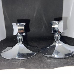 May include: Two shiny, chrome-finished candle holders. Each has a geometric base and a faceted cup to hold a candle. The candle holders are displayed on a dark surface, reflecting light. These decorative pieces are suitable for home decor.