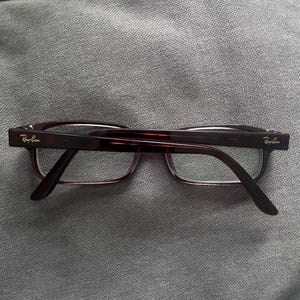 May include: A pair of Ray-Ban eyeglasses with a dark burgundy frame and temples. The lenses are clear, and the Ray-Ban logo is visible on the temples. The glasses are resting on a gray surface.