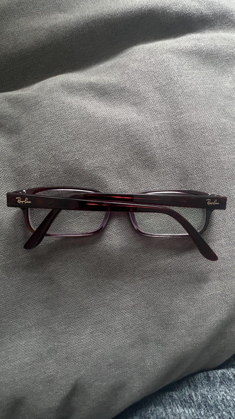 May include: A pair of Ray-Ban eyeglasses with a dark burgundy frame and temples. The lenses are clear, and the Ray-Ban logo is visible on the temples. The glasses are resting on a gray surface.
