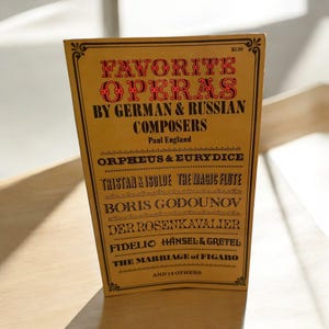 Puede incluir: Una cubierta de libro amarilla con el título "Favorite Operas by German & Russian Composers" de Paul England. La cubierta enumera óperas como "Orpheus & Eurydice", "Tristan & Isolde", "Boris Godunov", "Der Rosenkavalier", "Fidelio", "Hansel & Gretel" y "The Marriage of Figaro".