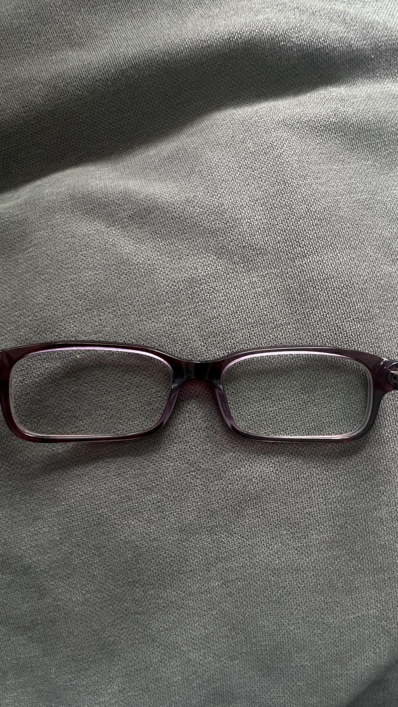 May include: Rectangular eyeglasses with a dark purple frame. The lenses are clear, and the glasses are set against a gray fabric background. The frames have a subtle, glossy finish.