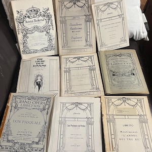 May include: A selection of antique opera librettos, including "Anna Bolena" and "Don Pasquale." The covers are mostly cream with black text and illustrations. The books are displayed on a dark brown surface.