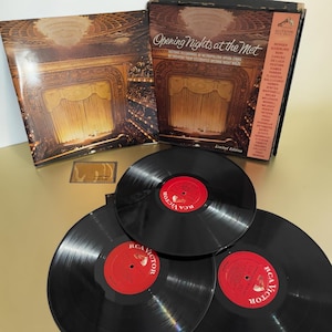May include: A vinyl record box set titled Opening Nights at the Met, featuring a brown cover with an image of an opera house interior. The set includes three black vinyl records with red RCA Victor labels and a small rectangular insert card.