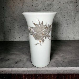 Puede incluir: Un jarrón de cerámica blanca con un diseño floral en tonos grises y dorados. El jarrón tiene una forma simple y elegante y es perfecto para exhibir flores.