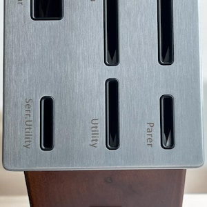 Puede incluir: Un bloque de cuchillos de acero inoxidable con ranuras para tijeras, Santoku, Chef, utilitario dentado, utilitario y cuchillos para pelar. El bloque tiene una base de madera marrón oscuro y una parte superior plateada.