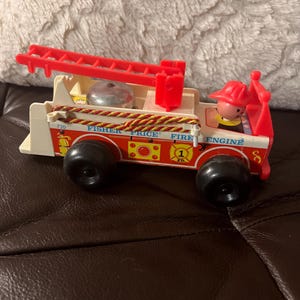 Könnte beinhalten: Ein Vintage-Spielzeug von Fisher-Price Fire Engine. Das Spielzeug ist rot, weiß und gelb mit schwarzen Rädern. Es verfügt über eine rote Leiter, eine silberne Glocke und eine Feuerwehrmannfigur. Der Text "Fisher Price Fire Engine" ist auf der Seite aufgedruckt.