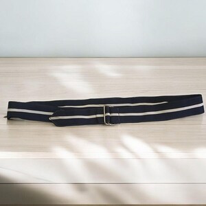 May include: A navy blue and white striped belt with a silver-colored buckle. The belt is lying flat on a light-colored wooden surface. The belt has two thin white stripes running lengthwise.