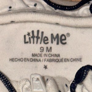 May include: Close-up of a white baby garment with a zipper. The garment has small dark blue polka dots and dark blue trim. The label reads "Little Me" and "9 M", with text indicating it was made in China.