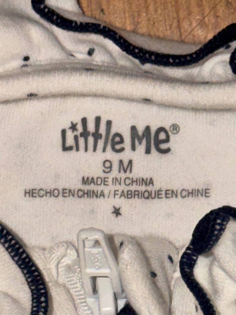 May include: Close-up of a white baby garment with a zipper. The garment has small dark blue polka dots and dark blue trim. The label reads "Little Me" and "9 M", with text indicating it was made in China.