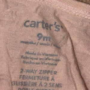 May include: Close-up of a pink Carter's baby garment label. The label indicates the size is 9 months and features text in English, French, and Spanish. The garment is made in Vietnam and has a 2-way zipper.