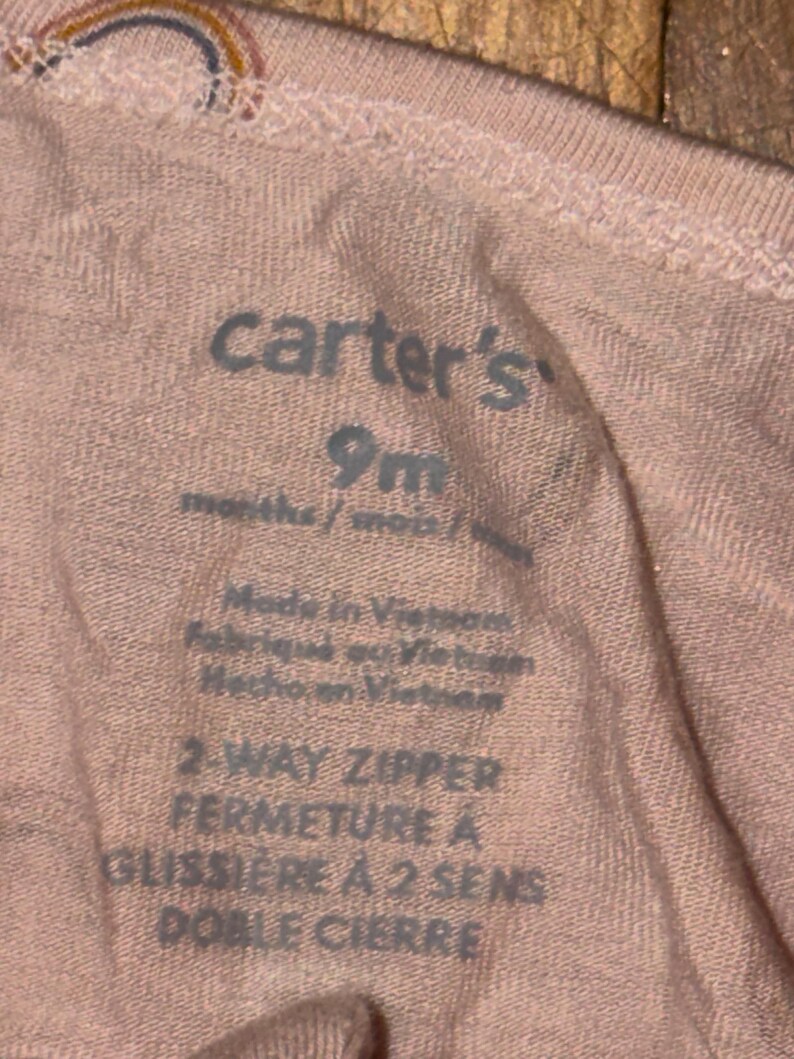 May include: Close-up of a pink Carter's baby garment label. The label indicates the size is 9 months and features text in English, French, and Spanish. The garment is made in Vietnam and has a 2-way zipper.