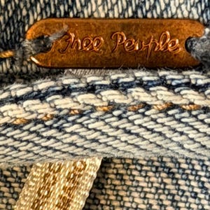 May include: Close-up of a copper-coloured tag with the words "Free People" embossed on it. The tag is attached to a denim fabric with a woven pattern of blue and white threads. A beige tag is visible.