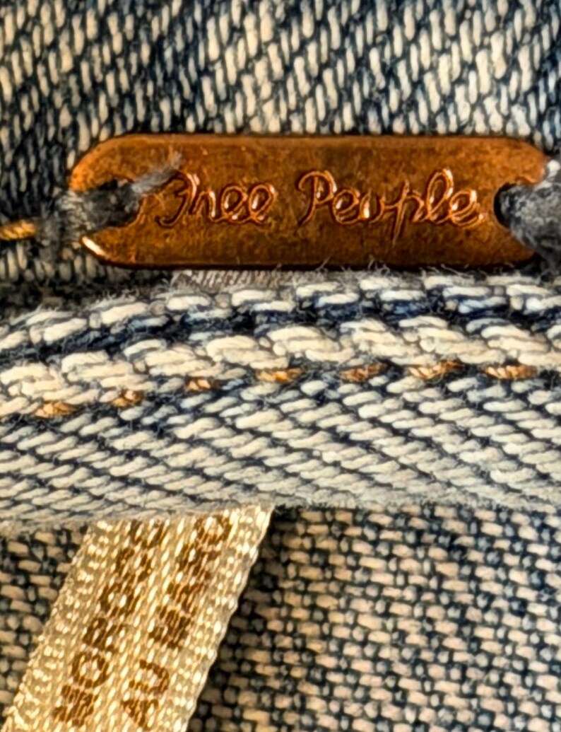 May include: Close-up of a copper-coloured tag with the words "Free People" embossed on it. The tag is attached to a denim fabric with a woven pattern of blue and white threads. A beige tag is visible.