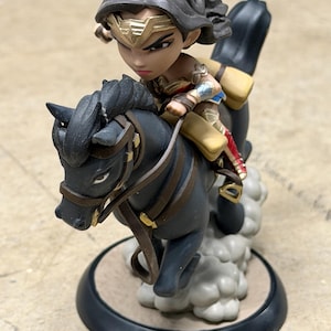 May include: A Wonder Woman figurine riding a black horse. The figure wears a gold tiara, armour, and a red and gold outfit. The horse has brown reins and is on a round base.