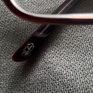 May include: Close-up of a pair of eyeglasses. The frame is a deep burgundy color with a rectangular shape. The Ray-Ban logo is visible on the temple. The glasses are set against a textured gray background.