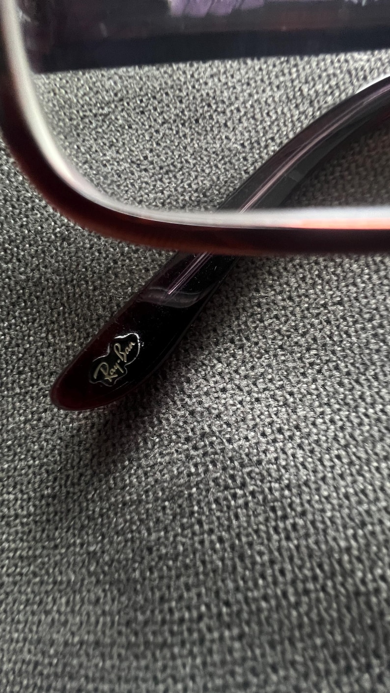 May include: Close-up of a pair of eyeglasses. The frame is a deep burgundy color with a rectangular shape. The Ray-Ban logo is visible on the temple. The glasses are set against a textured gray background.