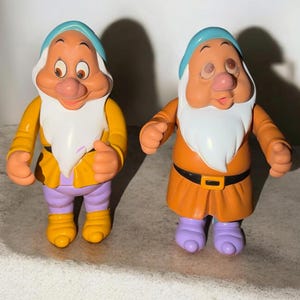 May include: Two plastic figurines of characters with large noses, white beards, and light blue hats. The left figure wears a yellow jacket and purple trousers, while the right figure wears an orange tunic with a black belt and purple shoes.