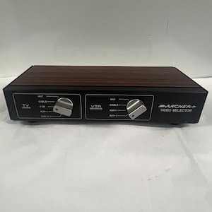 May include: A black Archer video selector with a woodgrain top. It has two sets of knobs for TV, VCR viewing and recording, and cable and antenna inputs. The text "ARCHER VIDEO SELECTOR" is printed on the front.