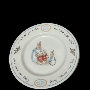 May include: A white ceramic plate with a blue rim features a depiction of Beatrix Potter's Peter Rabbit and his siblings. The plate has the text "I shall tell you a story about four little rabbits. Whose names were - Flopsy, Mopsy Cottontail and Peter." and "Peter Rabbit 1893-1993".