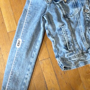 May include: Light blue denim jacket with a classic design. The sleeve length is 59.69 cm. Features include a button-up front, chest pockets, and a collar. The jacket is made of a light-wash denim material.