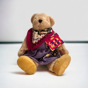 May include: A plush brown teddy bear wearing a purple dress and a pink and white patterned bandana. The bear has a yellow tag that says "Check Mates" with the Muffy logo.