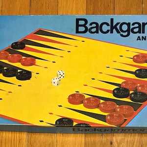 Puede incluir: Caja de juego de Backgammon vintage con un tablero amarillo y negro, fichas rojas y negras y dados. La caja azul muestra el título del juego y el logotipo de la empresa Milton Bradley.