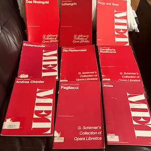 May include: A collection of red opera librettos from the G. Schirmer's Collection, featuring titles like "Das Rheingold," "Lohengrin," and "Porgy and Bess." Each book has white text and the word "MET" in large white letters.