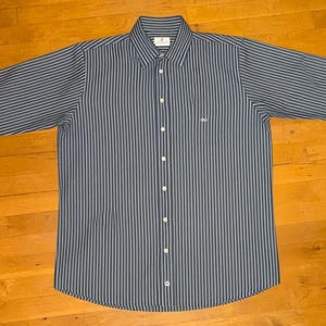 May include: A striped button-down shirt in blue and white, with long sleeves and a classic collar. The shirt features a small logo on the chest and is laid out flat. The shirt is displayed on a wooden surface.