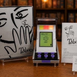 Könnte beinhalten: Electronic Taboo Platinum Edition Spielset bestehend aus einer silbernen Schachtel mit schwarzer Strichzeichnung, einem tragbaren elektronischen Spielgerät mit digitalem Bildschirm, der das Wort WEALTHY und Begriffe anzeigt, einer Anleitung und einer Infokarte.