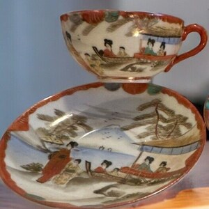 Peut inclure: Un service à thé vintage composé d'une tasse et d'une soucoupe. La tasse et la soucoupe sont décorées d'une scène peinte à la main de personnes dans des bateaux, des montagnes et des arbres. L'ensemble a une bordure rouge et orange.