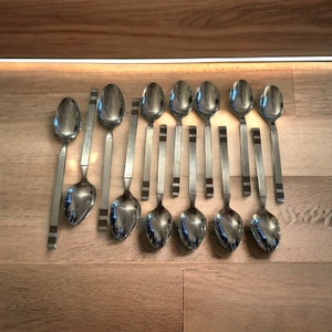 May include: A set of twelve stainless steel spoons arranged on a wooden surface. The spoons have a modern design with rectangular handles and a dark brown accent near the end. The spoon bowls are oval and reflective.