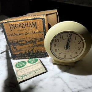 Puede incluir: Un despertador Ingraham vintage de color crema con un logotipo de Target, acompañado de su caja original e instrucciones de garantía. La esfera del reloj tiene números y manecillas negras. La caja dice "Clock Makers Over A Century."