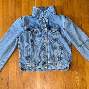 May include: A light blue denim jacket with a button-up front, collar, and long sleeves. The jacket has pockets on the chest and sides. The sleeves have a unique stitched design. The jacket is laid flat on a wooden surface.