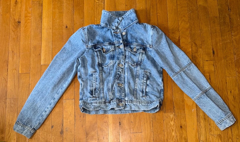 May include: A light blue denim jacket with a button-up front, collar, and long sleeves. The jacket has pockets on the chest and sides. The sleeves have a unique stitched design. The jacket is laid flat on a wooden surface.