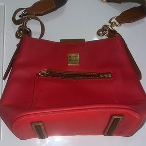 May include: Red leather crossbody bag with a brown zipper and a brown and olive green adjustable strap. The bag has a small front pocket and a gold-colored metal logo. The bag is a classic style.
