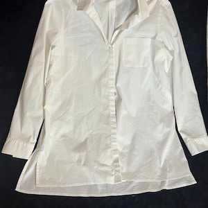 May include: A long-sleeved, white button-down shirt with a collared neckline and a single chest pocket. The shirt has a relaxed fit and a split hem design. The fabric appears to be a lightweight material, suitable for warmer weather.