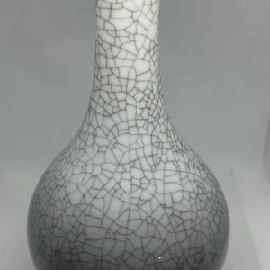 May include: A decorative ceramic vase with a crackle glaze pattern. The vase has a bulbous body that tapers to a slender neck and a flared rim. The base is dark blue with a gold rim. The vase is approximately 8 inches tall.